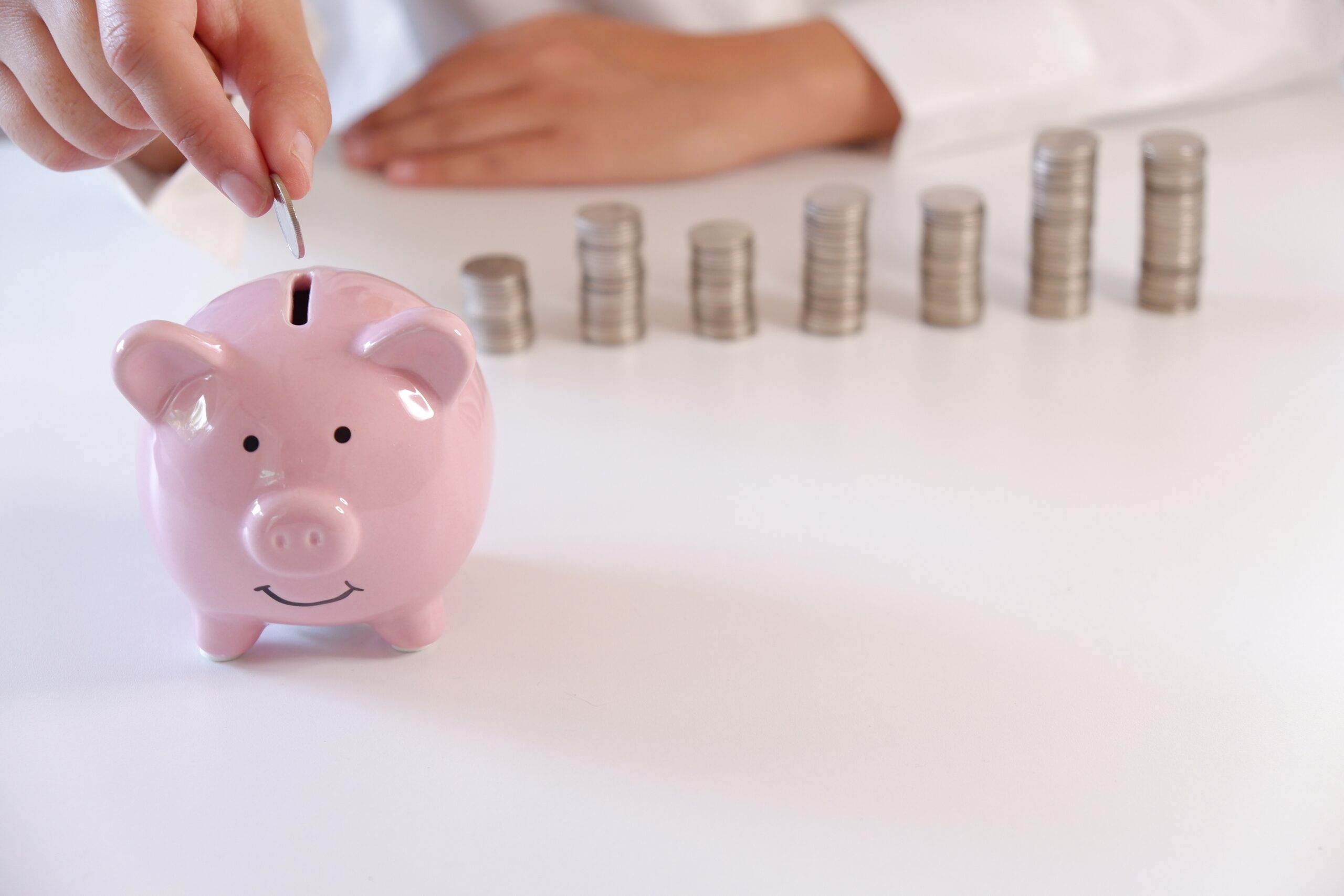 Businessperson Inserting Coins In Piggy Bank With Stack Of Coins Over The Desk